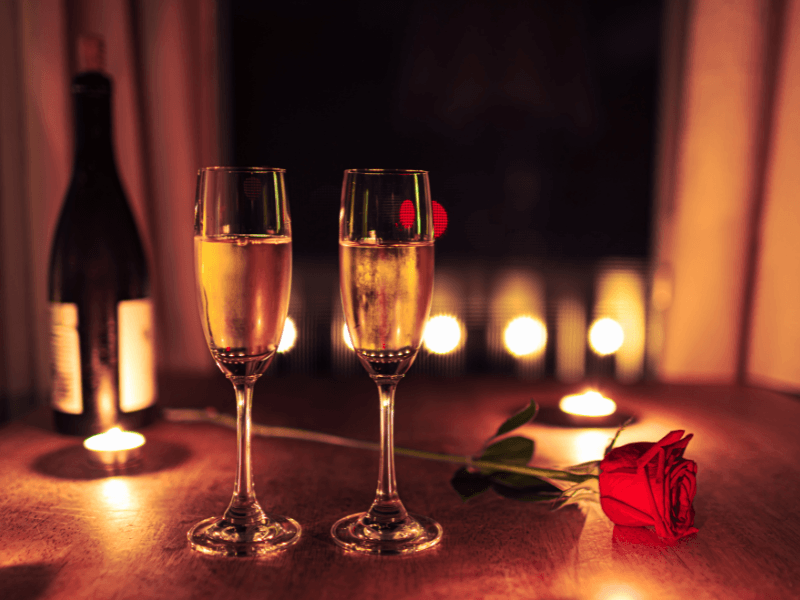 Twee gevulde champagneglazen op een houten tafel met een rode roos en brandende kaarsjes, op de achtergrond een fles wijn en zachte sfeerverlichting voor een romantische setting.