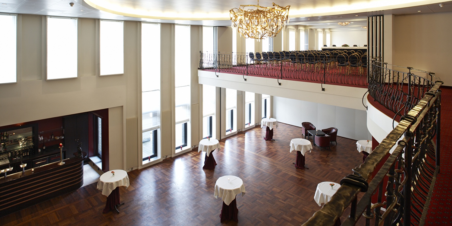 Geräumiger Ballsaal mit hohen Fenstern, Holzboden, Stehtischen mit weißen Decken, Balkon mit Metallgeländern und einem großen Kronleuchter.