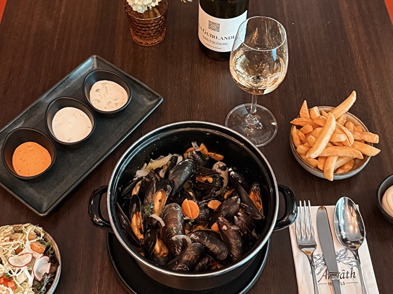 Geserveerde pan met verse mosselen in een zwarte pan, drie sausjes, friet, salade en een glas witte wijn (La Guirlande Sauvignon Blanc) op een donkerhouten tafel bij Amrâth Hotels.