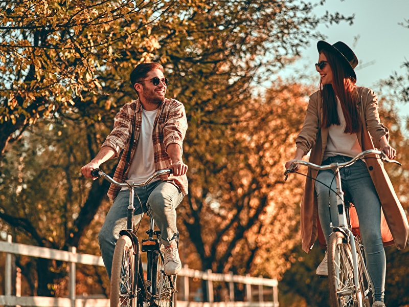 A man and woman are cycling next to each other, laughing, on a sunny autumn day.