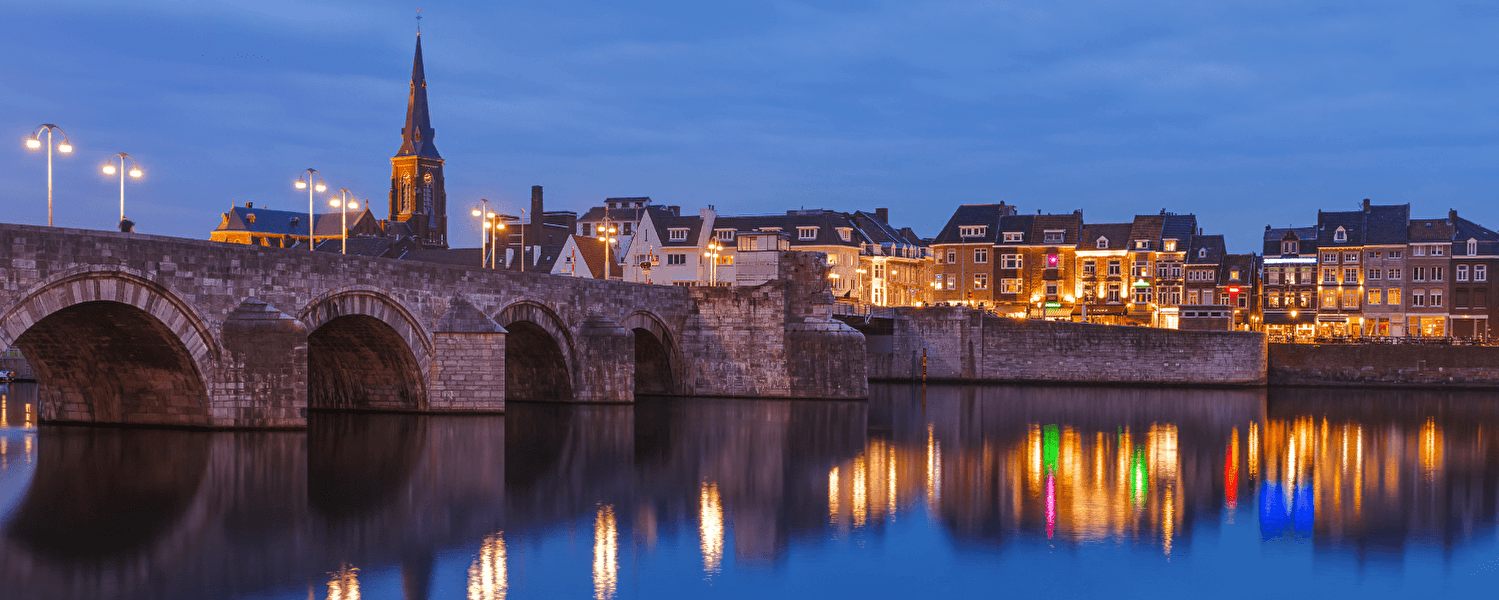 Abendblick auf die Sankt Servatiusbrücke und die beleuchtete historische Altstadt von Maastricht, mit Spiegelungen in der Maas.