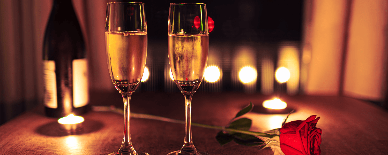 Zwei gefüllte Champagnergläser auf einem Holztisch mit einer roten Rose und brennenden Kerzen, im Hintergrund eine Weinflasche und sanfte Stimmungsbeleuchtung für eine romantische Atmosphäre.