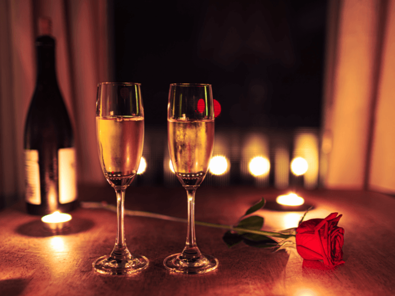 Twee gevulde champagneglazen op een houten tafel met een rode roos en brandende kaarsjes, op de achtergrond een fles wijn en zachte sfeerverlichting voor een romantische setting.